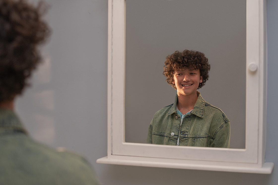 Image of a young boy with curly brown hair wearing a green shirt, smiling as he stands in front of a mirror